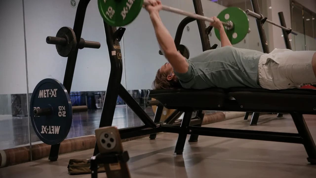 A member mid-set on a bench press at a gym, phone on a stand recording the set for the coach on the call